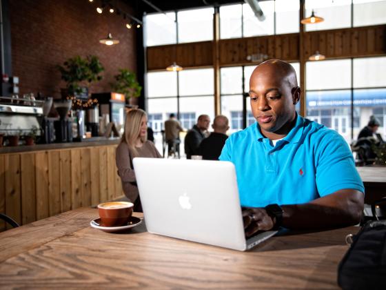 A black male student works on a laptop while sitting in a coffeeshop.