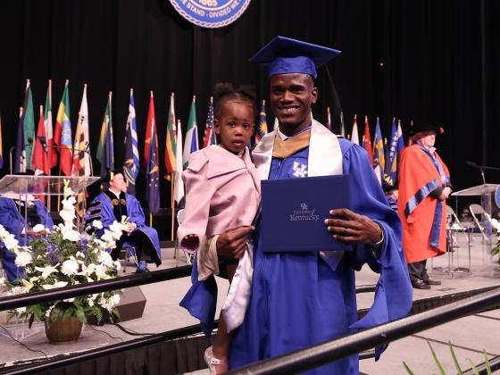 Student walking across stage holding diploma and child