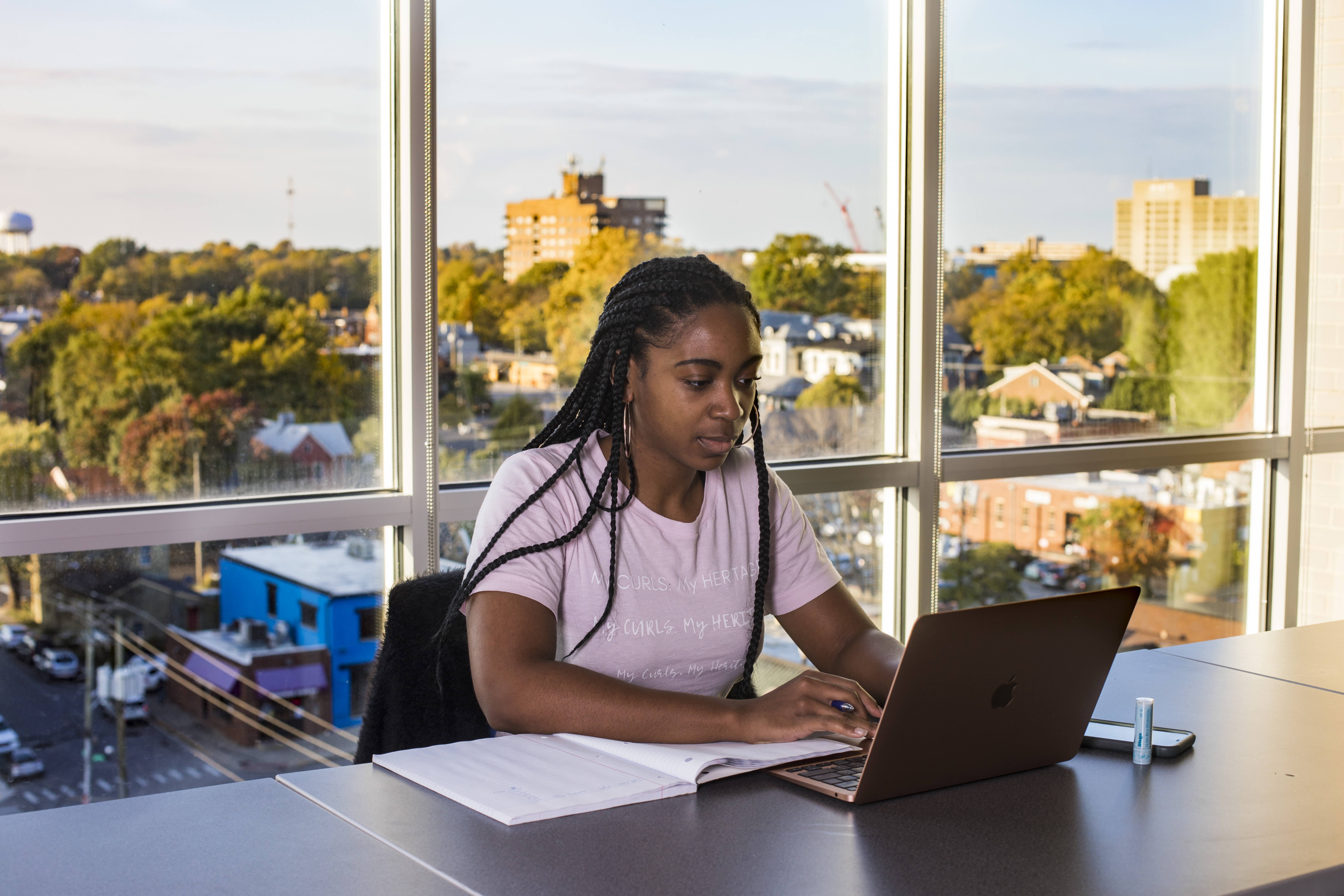 A black female student works at a laptop in her residence hall in front of a large window of campus.