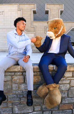 A young man in business attire sits on a stone wall and fist-bumps the UK Wildcat mascot, who is dressed in a blue suit outside the student center.