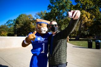 A white male student in a facial covering poses outside for a selfie with Wildcat.