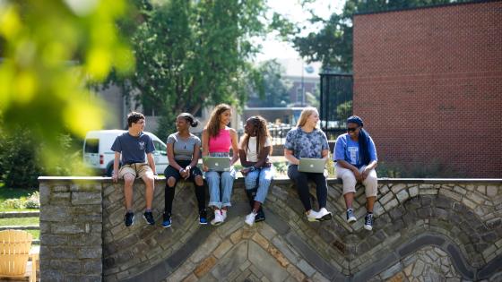 A group of six students sitting on a stone wall on the University of Kentucky campus, engaged in conversation and studies. The background features lush green trees and a clear blue sky, highlighting the vibrant and welcoming environment of the university.